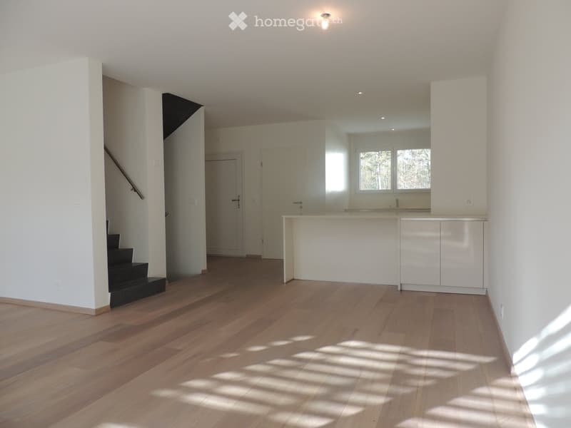 Empty room with wooden flooring, white walls, and a small kitchen area visible in the background. No furniture or equipment is present.