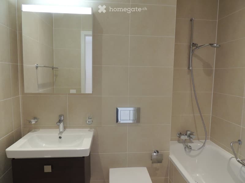 Modern bathroom with beige tiled walls, a white sink with a chrome faucet, a large mirror with built-in lighting, a bathtub with a handheld showerhead, and a toilet.