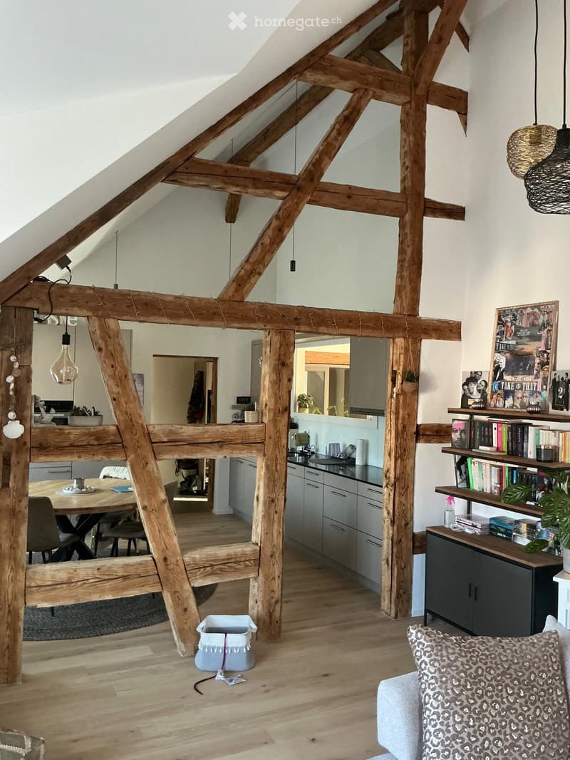 Open-plan living and dining area with exposed wooden beams, modern kitchen, and cozy seating area. Features include a sofa, dining table, pendant lights, and shelves with books and decor.