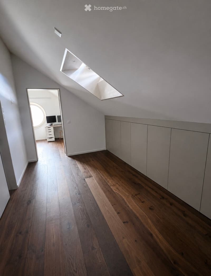 Bright, narrow hallway with wooden flooring, white walls, built-in cabinets on one side, and a skylight above. An open doorway leads to an adjacent room with a desk and computer monitors.
