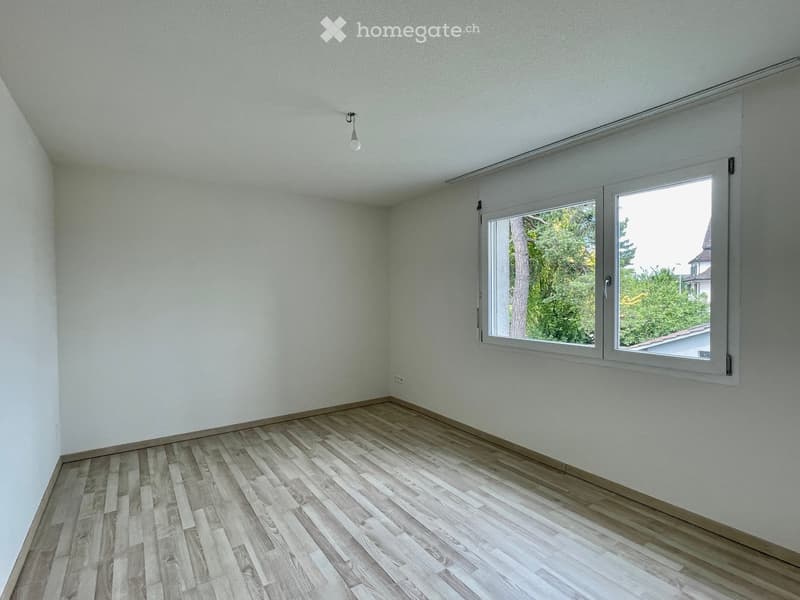 Empty room with light-colored walls, wooden flooring, and large windows with blinds.