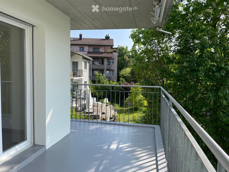 Balcony with metal railings, overlooking a garden, trees, and a distant building.