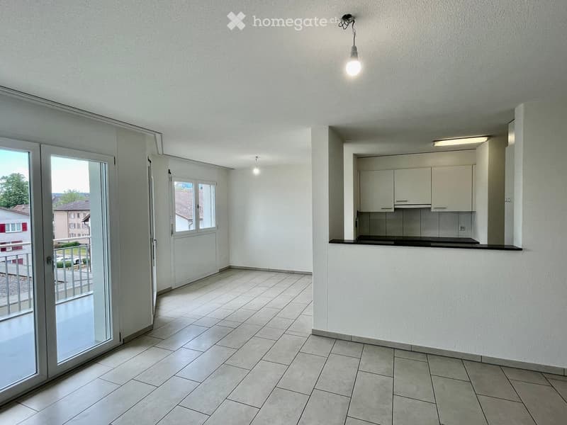 Empty room with tiled floor, white walls, and a small kitchen area with cabinets and a countertop. Features large windows and a sliding glass door leading to a balcony.