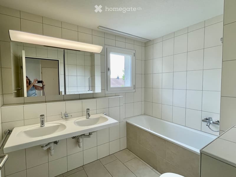 Bathroom with tiled walls and floor, double sink, bathtub, large mirror with built-in lighting, and a window providing natural light.