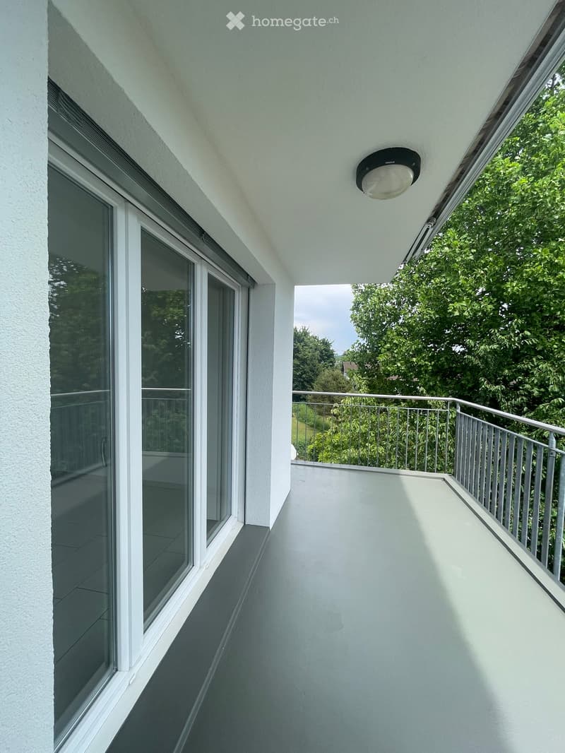 Balcony with white walls, grey tiled floor, metal railing, large windows, and a view of greenery.