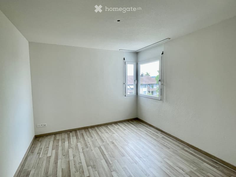 Empty room with light-colored walls and wooden flooring, featuring two windows with white frames and a visible electrical outlet on the left wall.