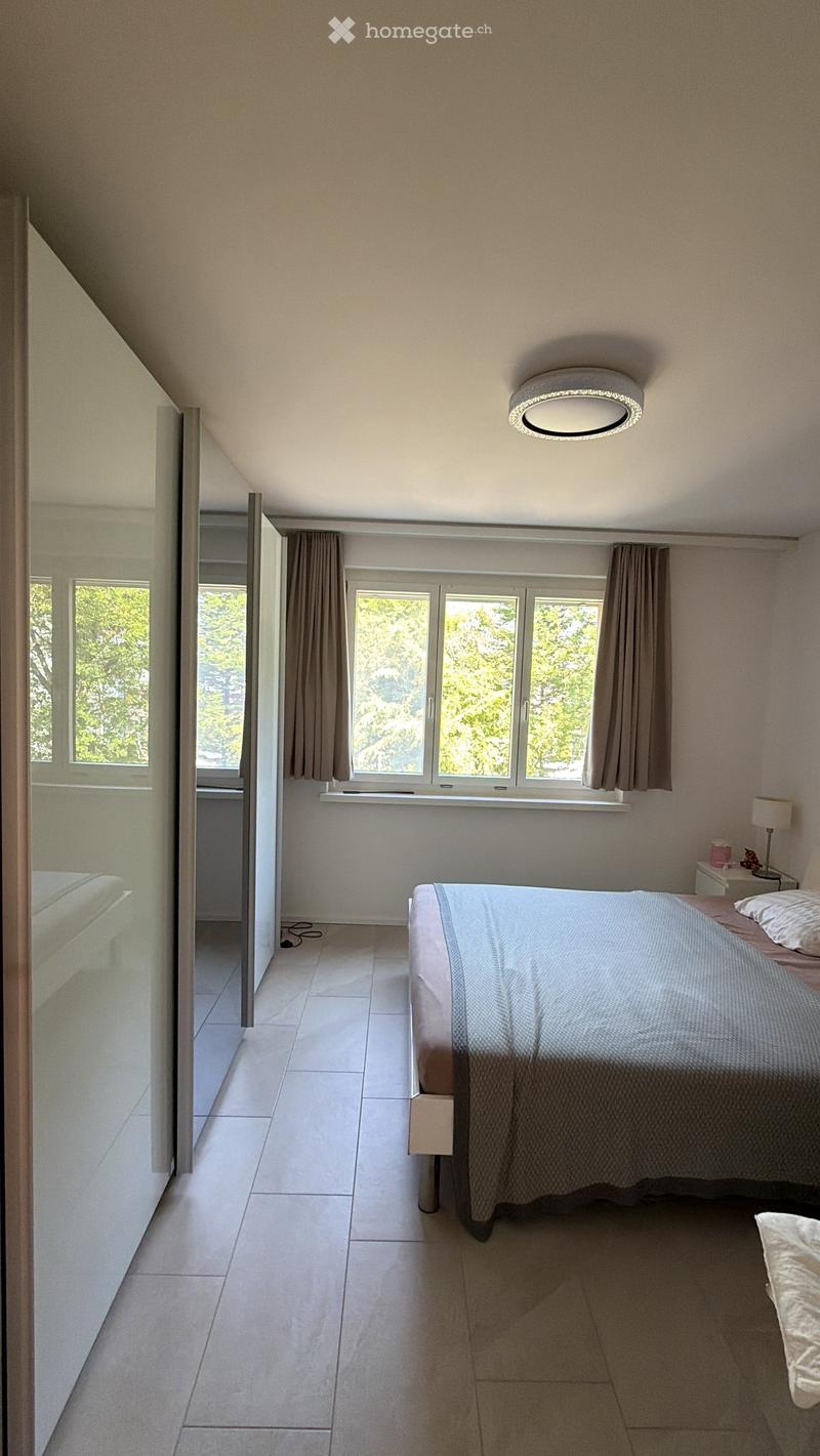 Modern bedroom with a large bed, nightstand, lamp, and large windows with curtains. The room features a sleek, minimalist design with a skylight ceiling fixture.
