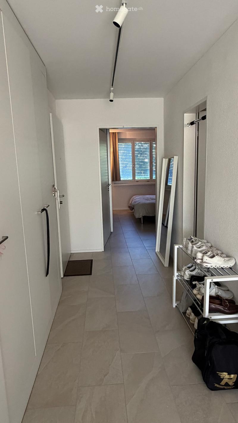 A clean, modern hallway with tiled flooring, white walls, and a shoe rack on the right side. A door leads to a bedroom visible in the background.