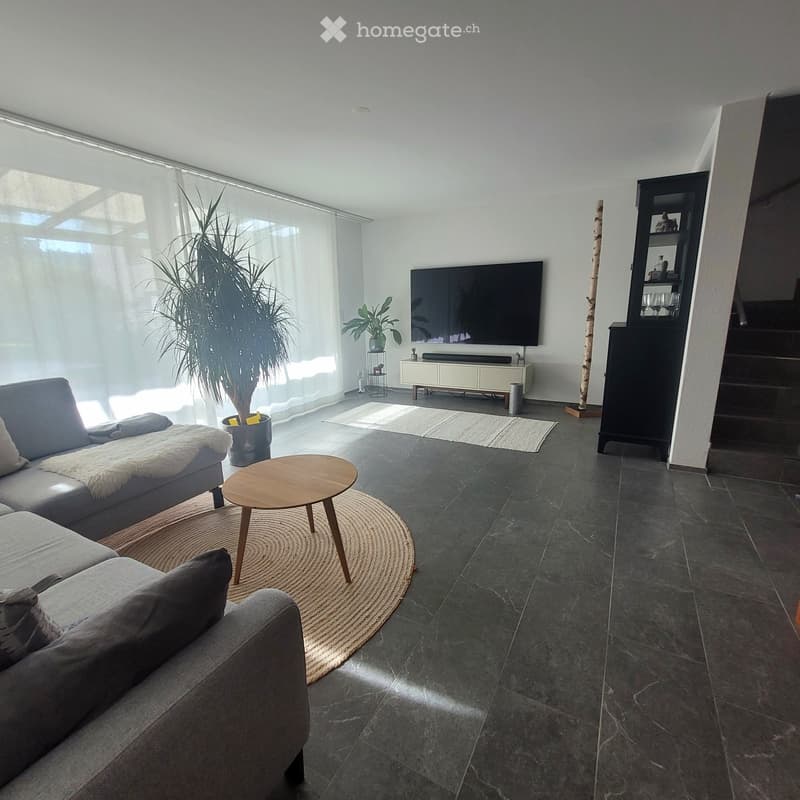 Modern living room with grey sectional sofa, wooden coffee table, large flat-screen TV, and indoor plants. The space features dark tiled flooring and large windows with sheer curtains.