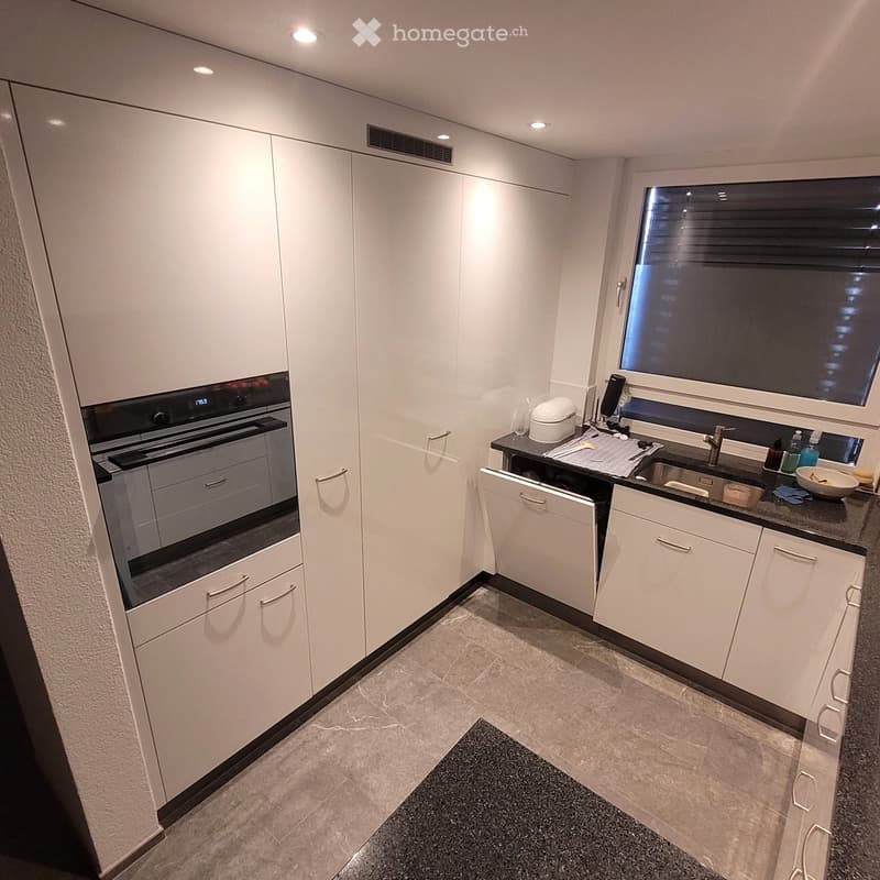 Modern kitchen with white cabinets, granite countertops, built-in oven, dishwasher, and sink area with various small appliances and dishes.