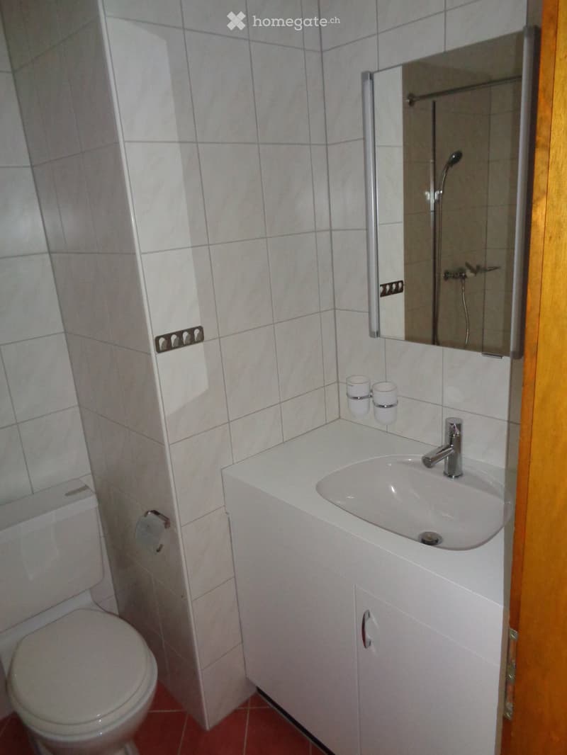 A small bathroom with a white sink, mirror, and toilet. The sink has a modern faucet and a small cabinet underneath. There are two cups on the sink counter and a wooden door frame visible on the right side.