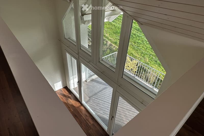 A modern, open corridor with wooden flooring and large windows leading to a balcony area with a view of greenery.