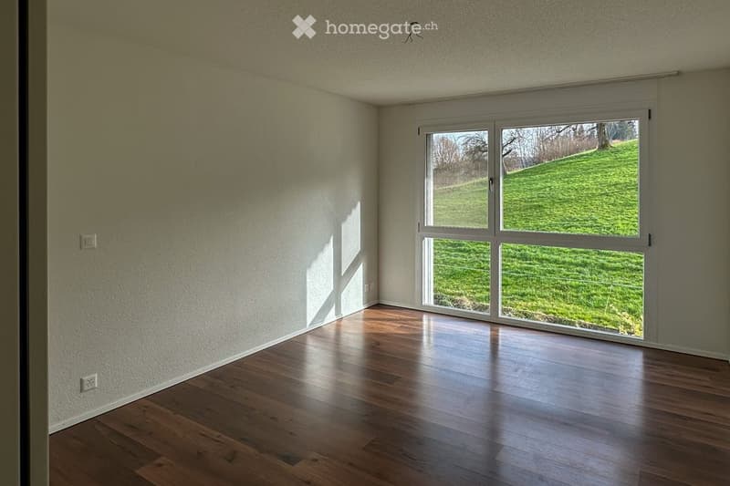 Empty room with wooden flooring, large window showing a green outdoor view, and white walls.