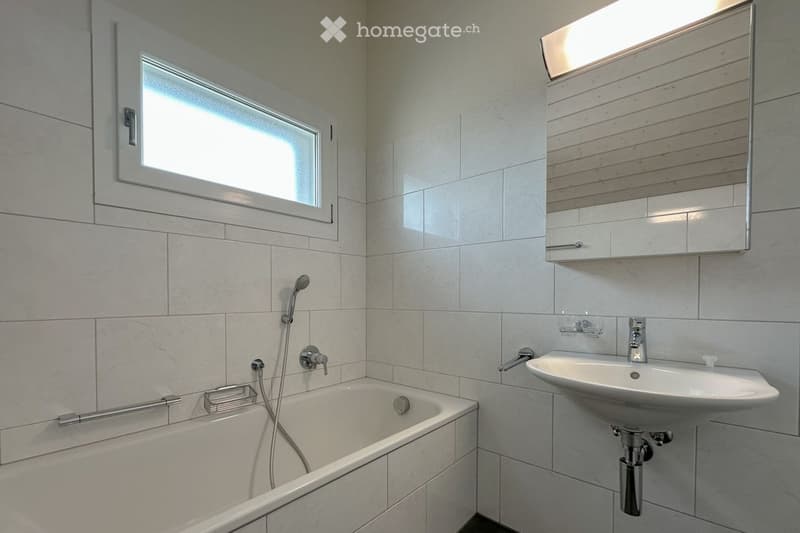 Modern bathroom with a bathtub, sink, mirror, and showerhead. The walls are tiled, and there is a window providing natural light.