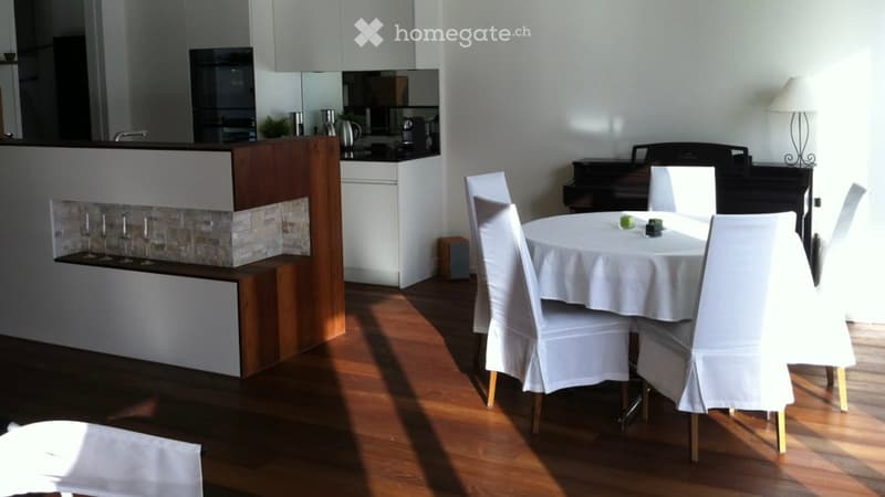 A modern dining area with a white tablecloth-covered table, six chairs, a piano, and a small kitchen area with cabinets and a faucet.