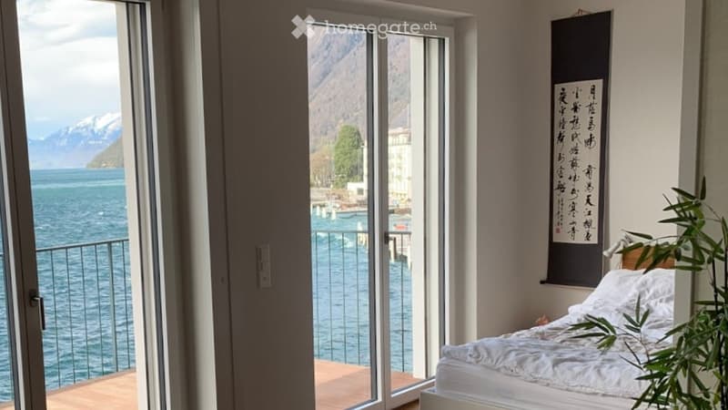 Bright bedroom with large windows offering a scenic lake and mountain view, featuring a neatly made bed and a potted plant near the window.