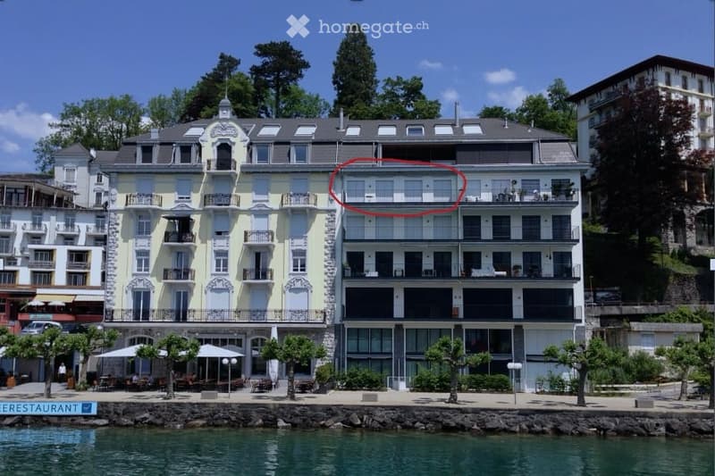 Multi-story building with balconies overlooking a waterfront, surrounded by trees and a restaurant area.