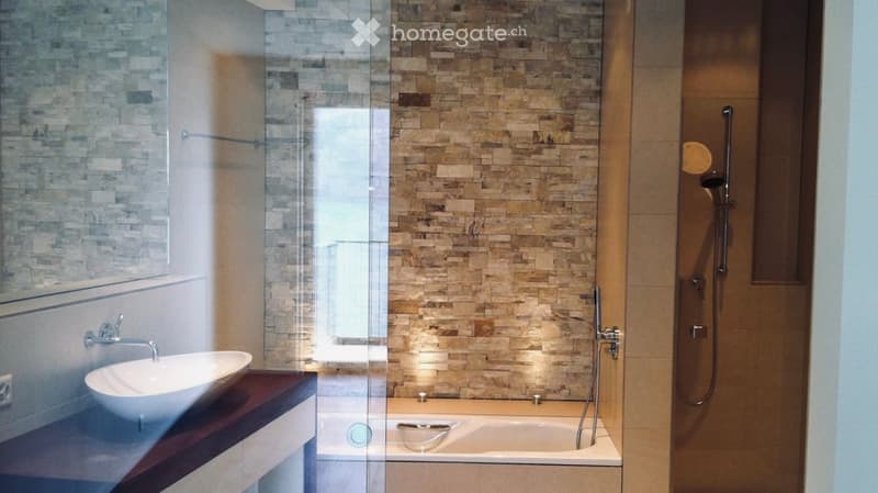 Modern bathroom with a sleek sink, bathtub, and glass-enclosed shower area. The walls are tiled in a stone-like pattern, and the space is well-lit with ambient lighting.