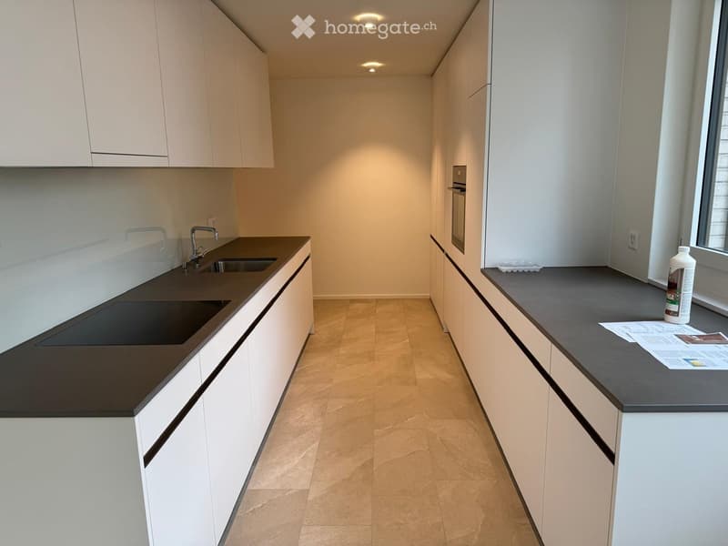Modern kitchen with white cabinets, dark countertops, and a built-in sink and oven. A bottle is placed on the counter near the window.