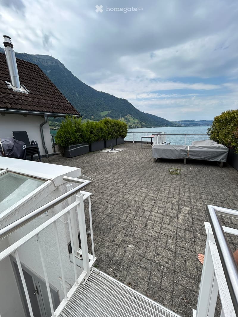 Roof terrace with stone tiles, white metal railings, covered sofa, chairs, and a view of a lake and mountains.