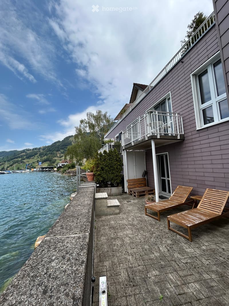 Balcony overlooking a lake with wooden lounge chairs, a small table, and a bench near the entrance.