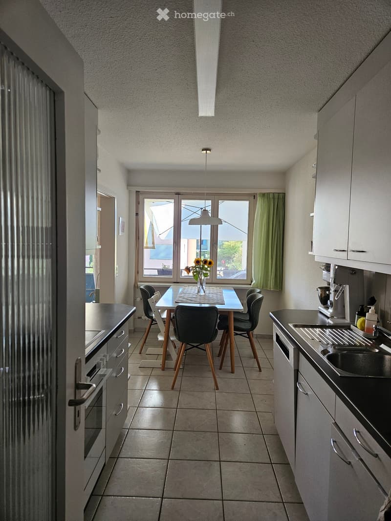 Modern kitchen with white cabinets, black countertops, a sink, oven, and a small dining area with a table and chairs. A vase with sunflowers sits on the table, and a window with green curtains lets in natural light.