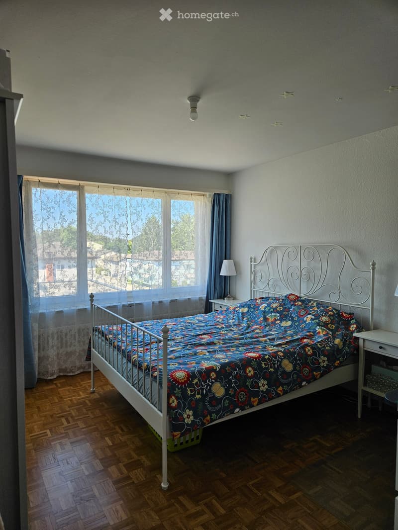 Bedroom with wooden floor, large window with curtains, white metal bed frame, colorful bedspread, nightstand with lamp.