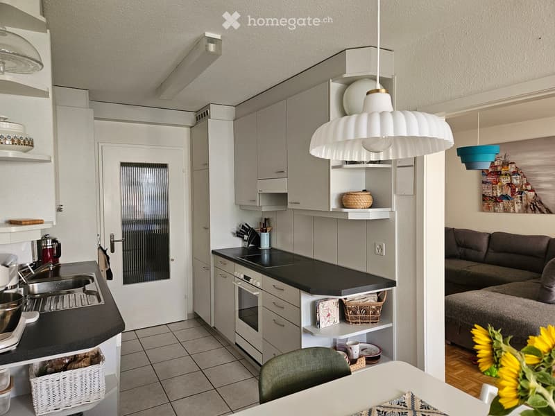 Modern open-plan living and kitchen area with white cabinets, grey sofa, dining table, pendant lights, and decorative items.