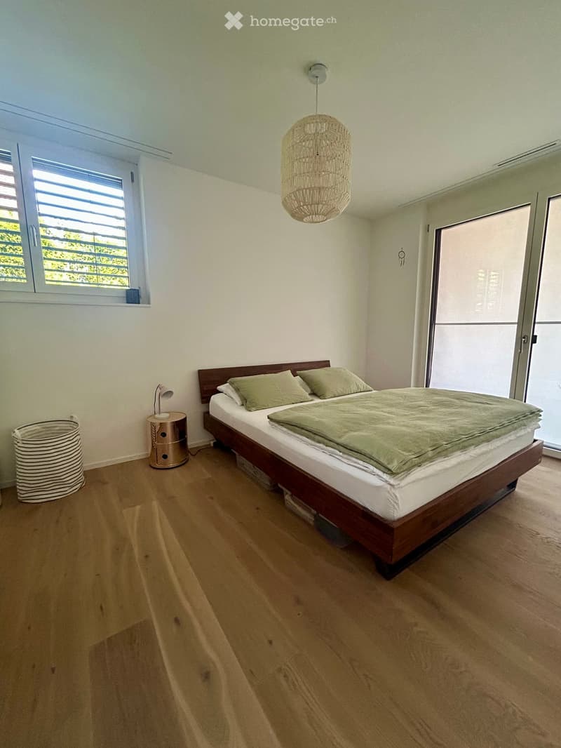 Modern bedroom with wooden flooring, a large bed with green bedding, a nightstand with a lamp, and a wicker basket near the window.