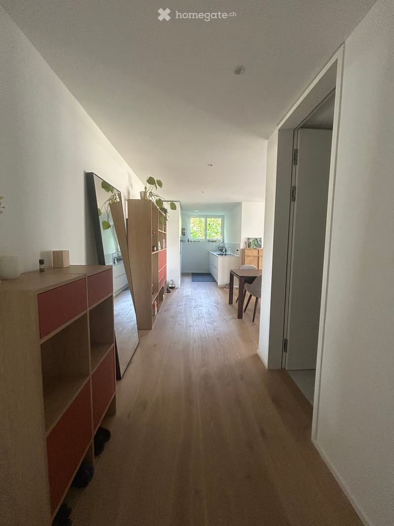 A long, narrow hallway with wooden flooring and white walls, leading to a kitchen area with a dining table and chairs, and a shelf with decorative items on the left side.