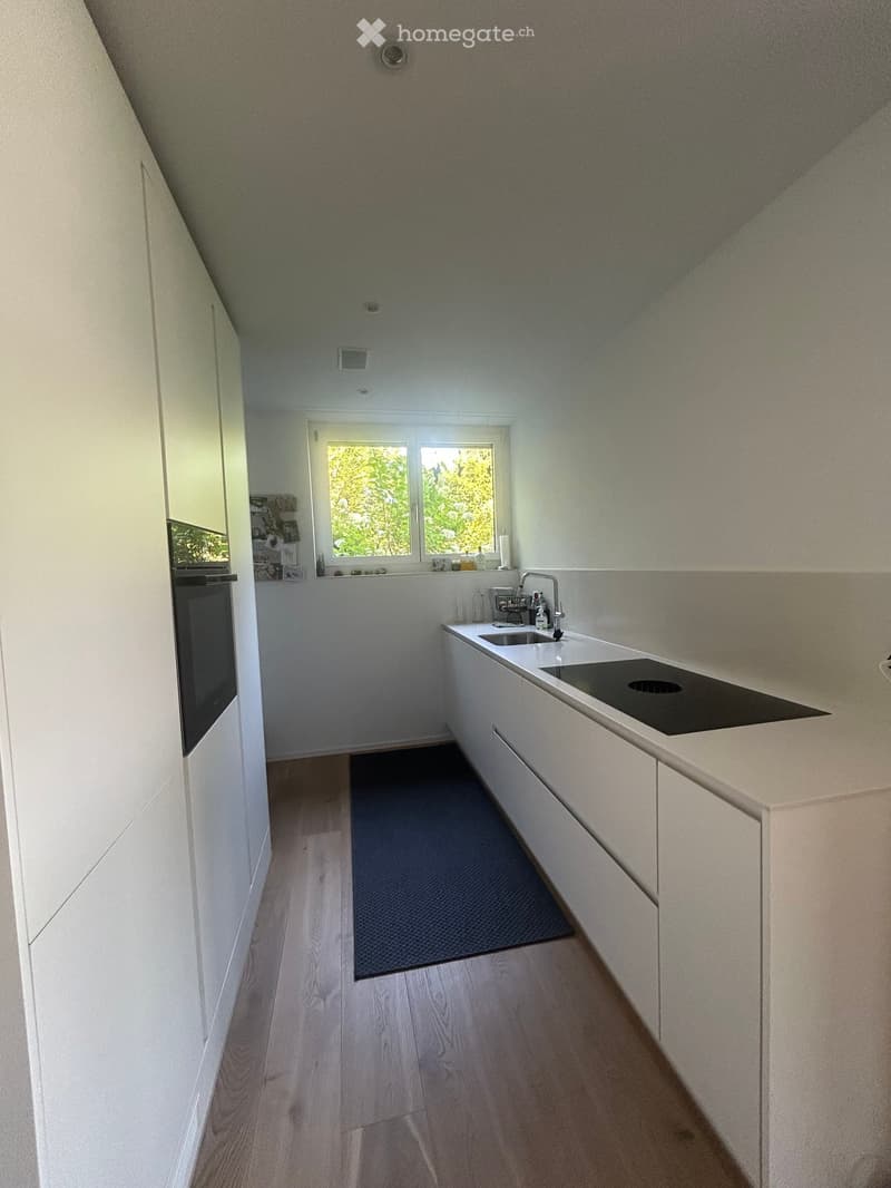Modern kitchen with white cabinets, light wooden flooring, and a sleek countertop featuring a sink and faucet. A dark-colored mat is placed on the floor.