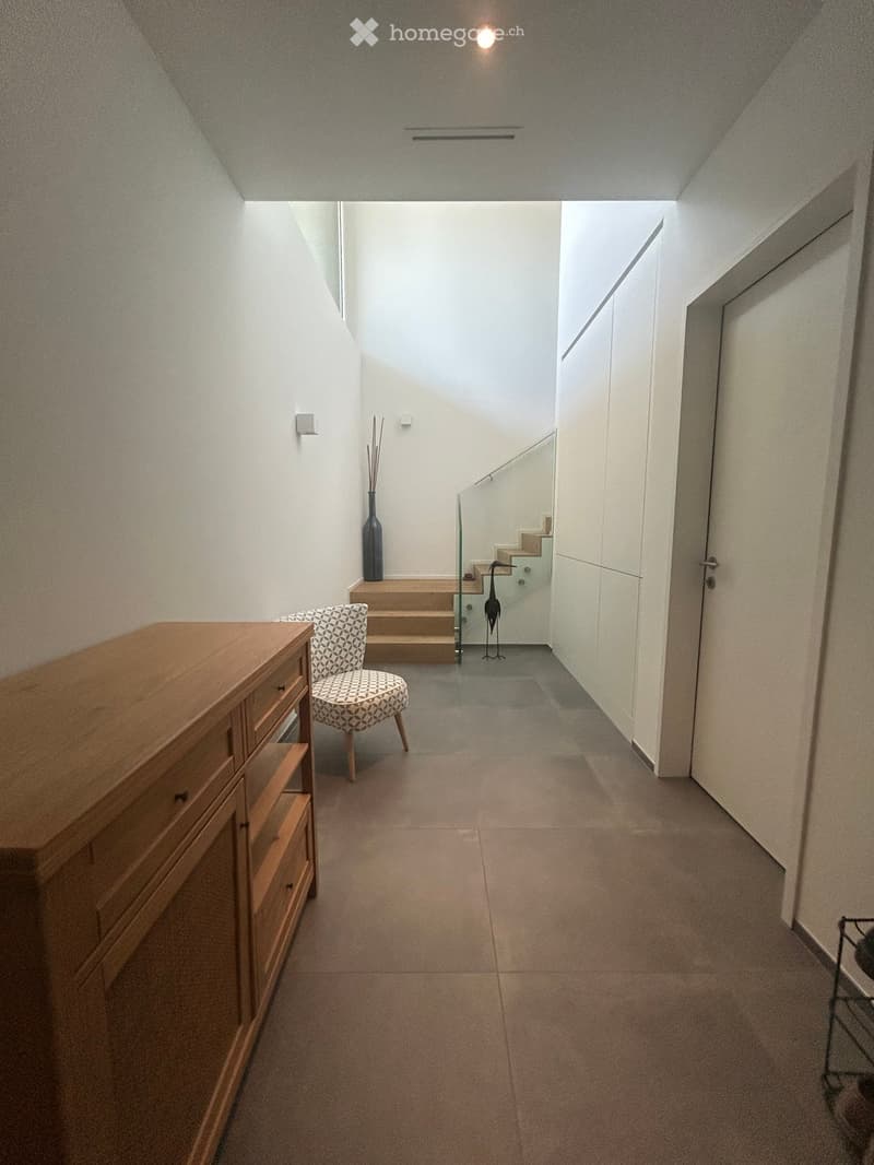 Modern hallway with tiled floor, white walls, wooden cabinet, patterned chair, decorative vase, and glass staircase leading upwards.