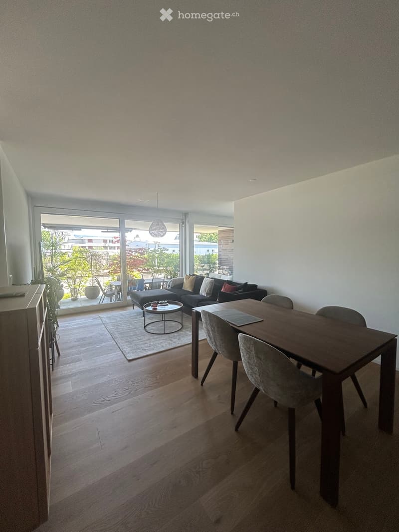 Modern living and dining area with wooden flooring, large windows, and a balcony view. Features a sofa, coffee table, dining table, and chairs.
