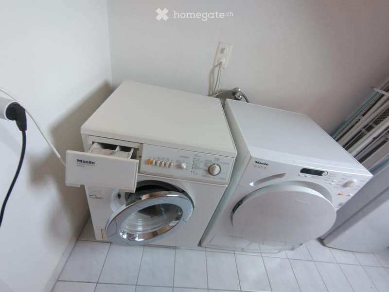 A small bathroom with a white washing machine and a white dishwasher placed side by side against a tiled wall.