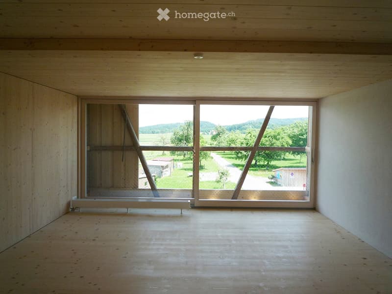 Empty room with wooden walls and ceiling, large windows showing a rural landscape view, and a simple wooden floor.