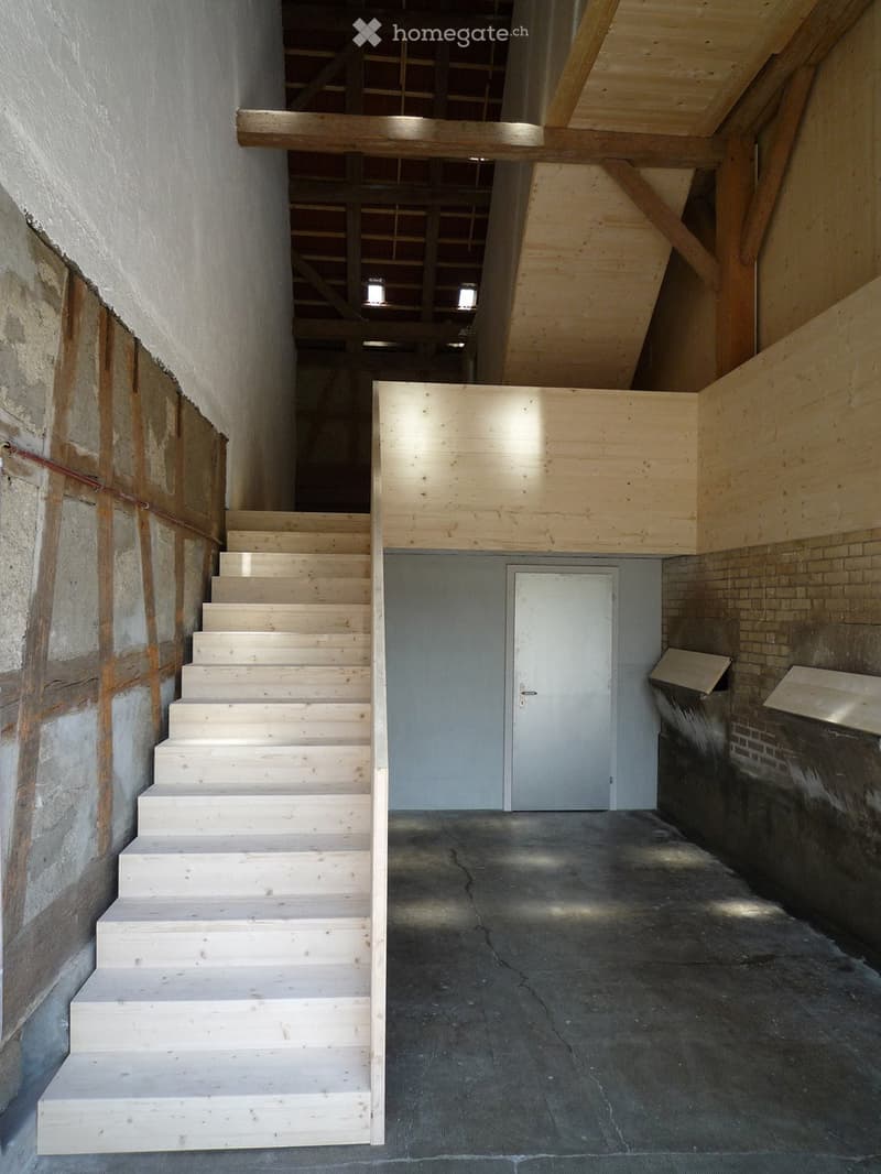 Staircase leading to an upper level with exposed wooden beams and a concrete floor. The walls are partially unfinished with visible brickwork and plaster.