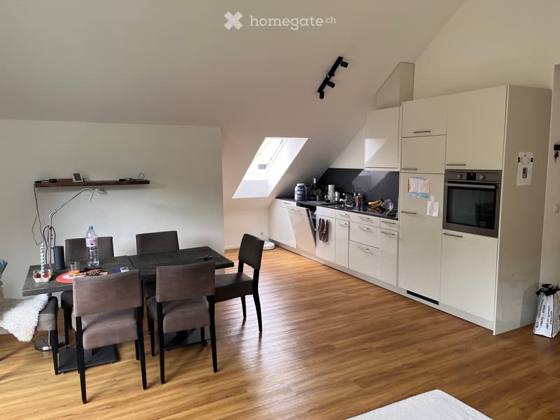 A modern, compact kitchen and dining area with white cabinets, a black dining table, four chairs, and various kitchen appliances including an oven, dishwasher, and refrigerator. The space features wooden flooring and a skylight window providing natural li