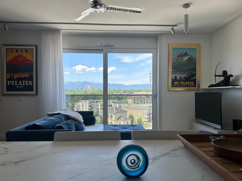 Living room with white walls, sliding glass door with city view, blue sofa, flatscreen TV, table with decorative item, two framed pictures, ceiling fan, white curtains, wooden side table
