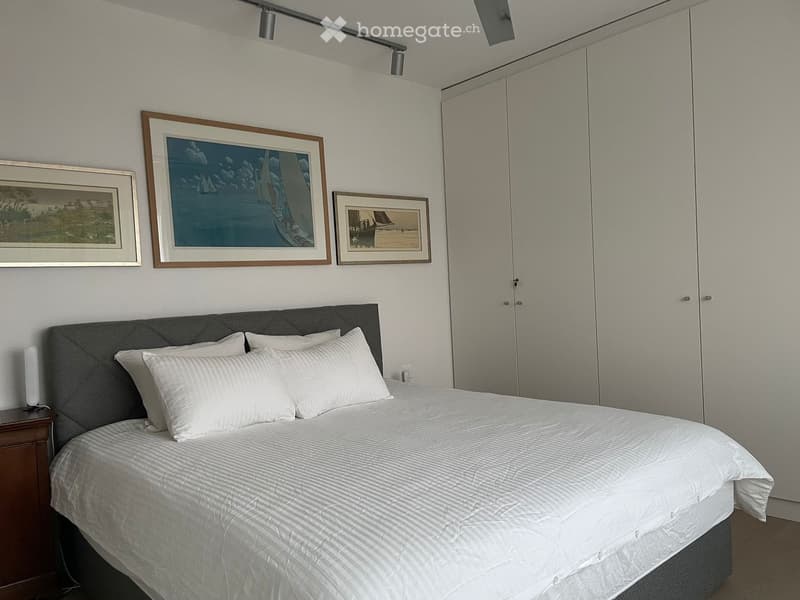 Queen size bed with grey headboard, white bed linen, white duvet, 3 white pillows, bedside table with lamp, built-in wardrobe, wall with 3 framed paintings, white walls, ceiling lights