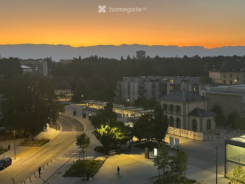 Aerial view of city, multiple buildings, streetlights, trees, people walking, mountains in the distance, sunset sky, and cars parked.