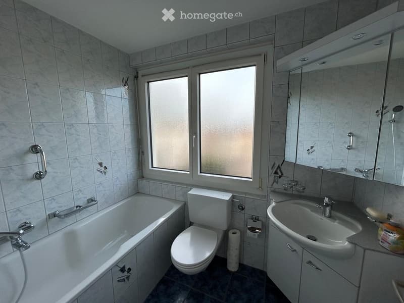 Bathroom with a bathtub, toilet, sink, mirror, and cabinet. The room features tiled walls and a window with frosted glass.