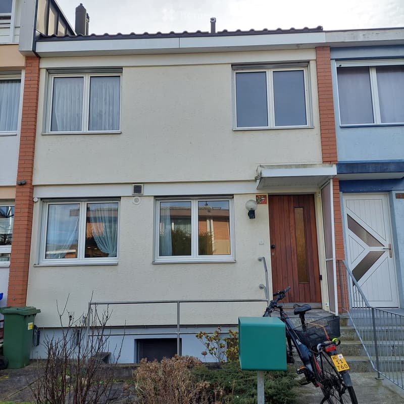 Two-story house with white siding, brick accents, multiple windows, and a wooden front door. A bicycle is parked near the entrance, and there is a green trash bin to the left.