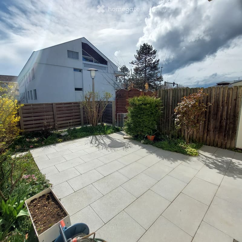 Garden area with tiled patio, wooden fence, various plants, and a modern house in the background.