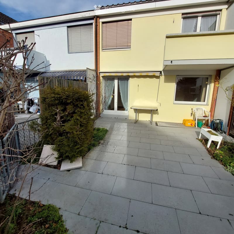 Patio area with tiled floor, small table, chairs, and greenery near a two-story house.