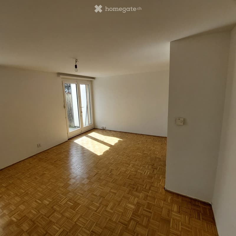 Empty room with wooden parquet flooring, white walls, and a large window with blinds allowing natural light to enter.