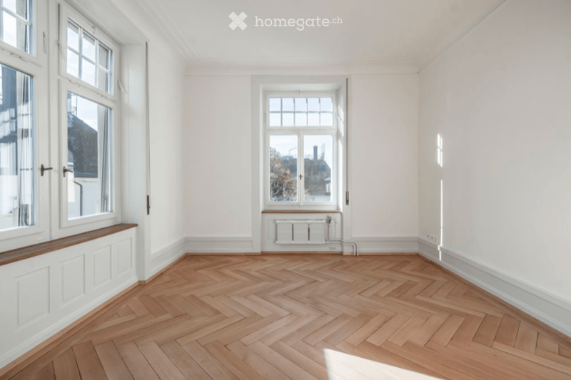 Completely empty room with white walls, herringbone wooden flooring, and large windows allowing natural light. No furniture or equipment present.