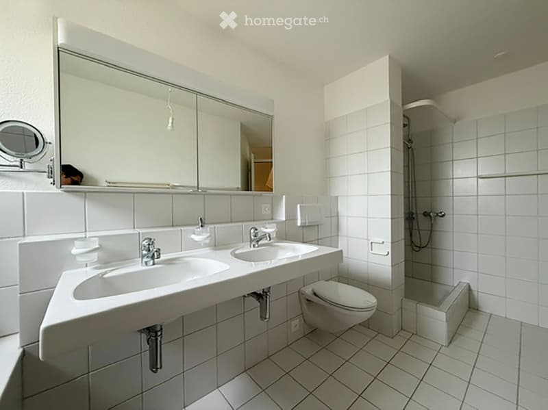 Modern bathroom with white tiles, double sink, large mirror, toilet, and shower area.