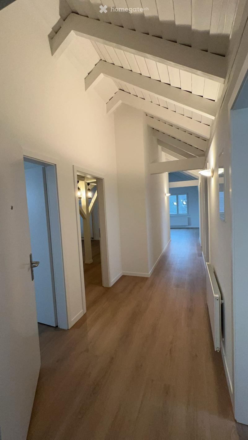 A modern hallway with wooden flooring, white walls, and exposed wooden beams on the ceiling. The hallway features several doors leading to different rooms and is illuminated by wall-mounted lights.