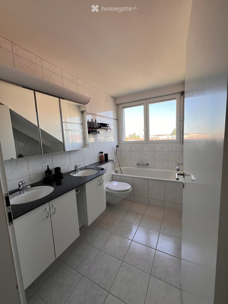 Modern bathroom with two sinks, a toilet, a bathtub, and a large mirror above the sinks. The room features light-colored tiles and a window providing natural light.