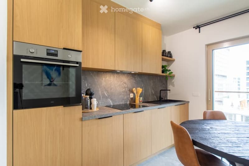 Modern kitchen with light wood cabinets, stainless steel oven, sink, and countertop. Includes a small dining area with a table and chairs.